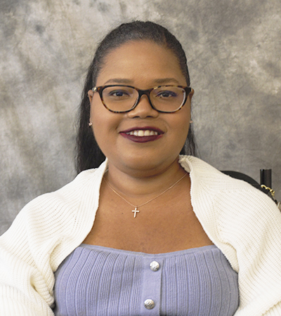 Woman sitting in a wheelchair with straight black hair wearing glasses and a white sweater over blue blouse