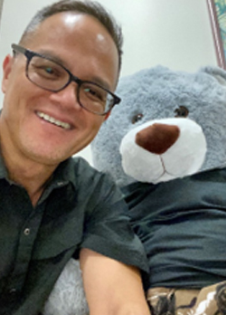Man with short grey hair, wearing black rimmed glasses and an olive green short sleeve button up, posing next to a teddy bear dressed similarly