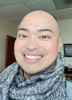 Man with shaved head smiling, wearing a grey patterned scarf